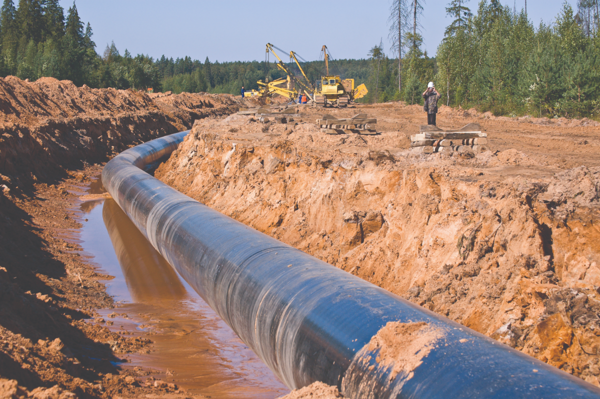 Photo d'une tranchée creusée dans le sol dans laquelle se trouve un tronçon de pipeline. Un ouvrier en uniforme se tient près de la tranchée et des véhicules de construction sont visibles au loin.
